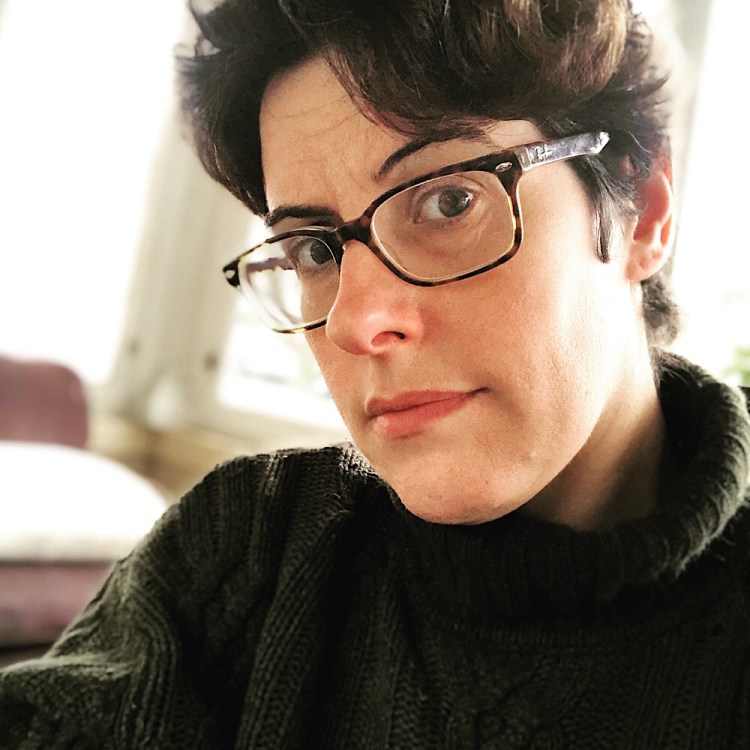 This image is a photograph of founder and publisher Angel Ackerman, a white woman with short, dark hair, big brown eyes and glasses